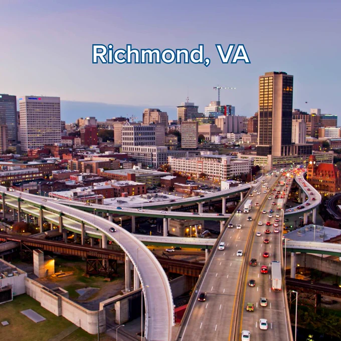 An aerial view of Richmond, Virginia. The image showcases a complex highway interchange with moving traffic in the foreground, leading toward the city's downtown skyline. The sky is a soft blend of blue and purple. The text "Richmond, VA" is centered at the top in white.