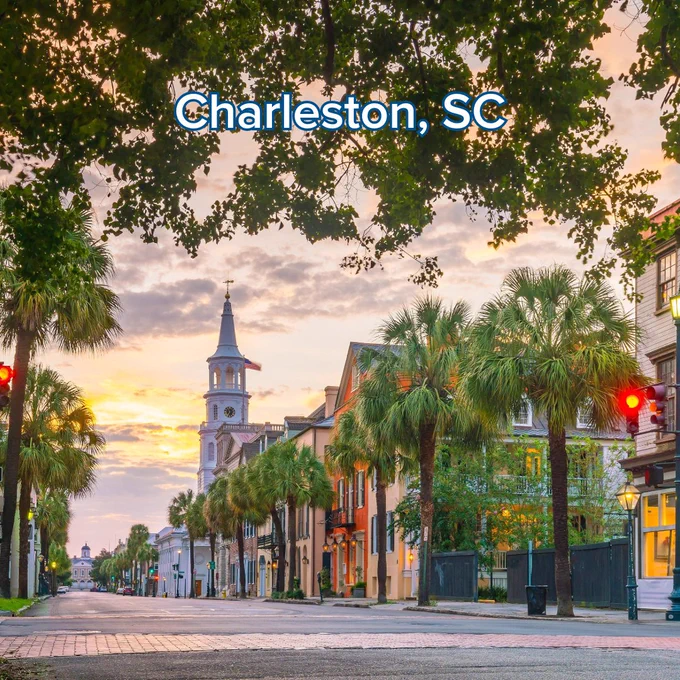 A scenic street view of Charleston, South Carolina, at sunset. The image shows historic buildings, vibrant green palm trees, and the prominent white steeple of St. Philip's Church in the distance. The sky is filled with warm, golden light. The text "Charleston, SC" is centered at the top.