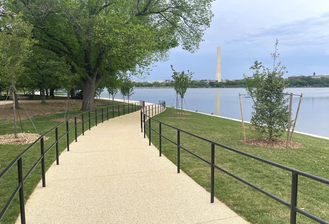 A concrete path running by grassy lawns and newly planted trees towards with the calm waters of the Tidal Basin on the right side and the Washington Monument in the background.