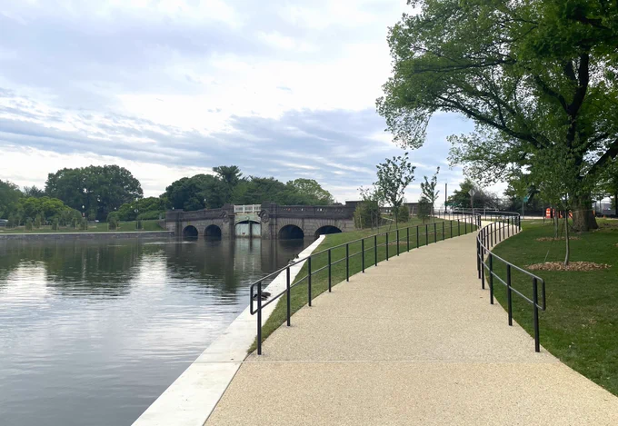A concrete path curving along the calm waters of the Tidal Basin and up a ramp bordered by grassy lawns and newly planted trees.