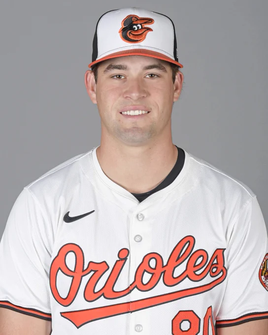 JT Quinn looks at the camera and smiles. He is wearing a white Orioles jersey and a tri-color cap.