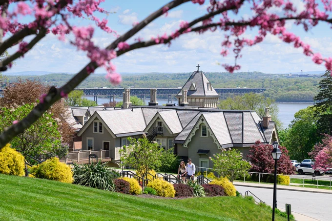 A pink flowering tree frames a springtime image of Mount Saint Mary College's campus. The Villa is seen with students walking up the stairs, and behind is the Newburgh-Beacon Bridge and Hudson River. 