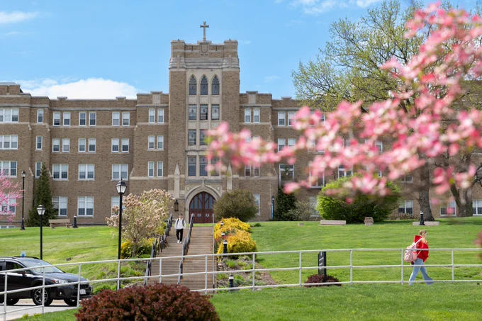 Springtime scene at Mount Saint Mary College. Students are walking around campus, the pink flowered tree in the foreground is blurred, and the Dominican Center is shown in the background. 