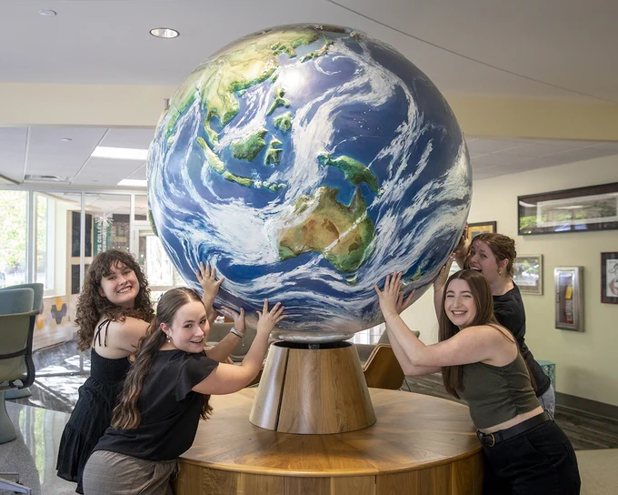 Four people are joyfully posing around a large globe, depicting Earth with vibrant blue and green colors, inside a well-lit room. Some framed artworks are visible on the walls in the background.