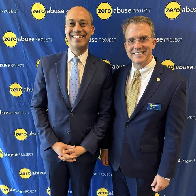 Two men in suits smile and pose together in front of a blue step and repeat banner printed with the Zero Abuse Project logo. The man on the right wears a name badge identifying him as Jeff Dion, Chief Executive Officer of the Zero Abuse Project.