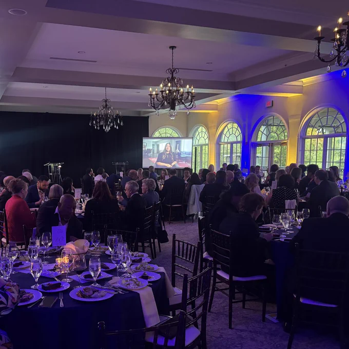 A wide shot of a formal gala dinner in a ballroom with chandeliers and arched windows. Guests are seated at round tables with navy linens and glassware. A projection screen at the front of the room displays a video, and blue and amber uplighting illuminates the walls.
