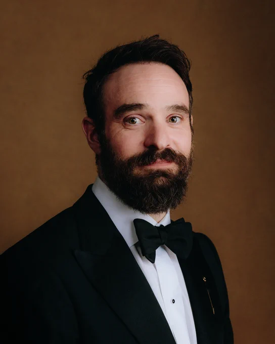 Chalie Cox smiles at the camera in a black tuxedo.