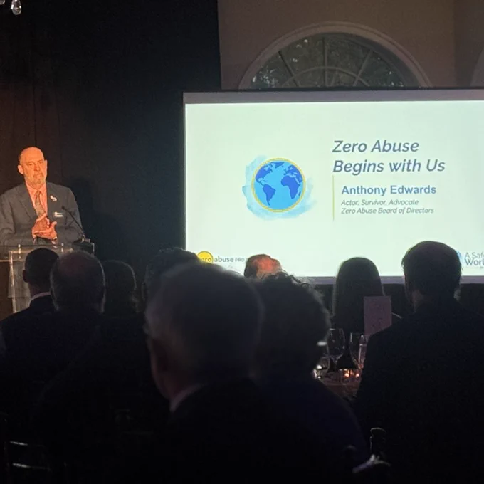 A speaker stands at a podium addressing a seated audience in a dimly lit event venue. A presentation screen behind him displays the slide "Zero Abuse Begins with Us" with the name Anthony Edwards listed as Actor, Survivor, Advocate and Zero Abuse Project Board of Directors.