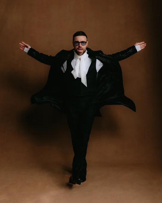 Ben Starr poses stepping forwards with his arms raised, having flicked his coat tails. His mouth is open and he is wearing a black suit and glasses.
