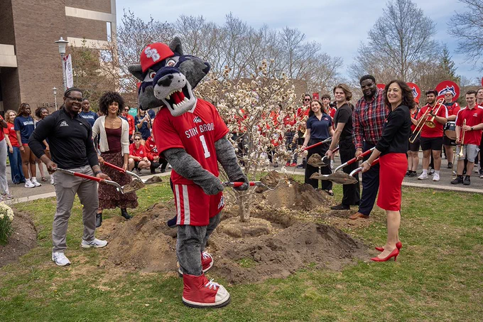 Wolfie, President Goldsmith and attendees stand around a newly planted tree while holding shovels during an inauguration week ceremony.