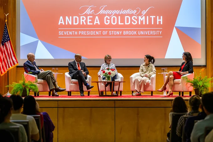 Panelists sit on stage during a discussion as a large presentation screen is displayed behind them.
