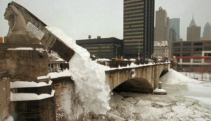 A bridge covered in snow with large chunks of snow cascading from a truck over the edge into a frozen river below. In the background, several tall buildings and skyscrapers are visible, partially obscured by a gray, overcast sky.