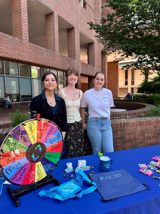 Three people stand behind a table with a colorful prize wheel and promotional items outdoors near a brick building.
