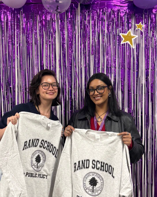 Two RAND School students pose with matching school sweatshirts in front of a sparkling purple backdrop and balloons. Two yellow stars are doodled on the graphic.