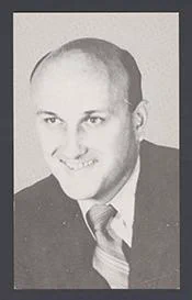 The image is a black and white photograph of a white man with balding hair wearing a suit and tie. He is smiling and appears to be posing for the picture. The man is wearing a black suit, white collared shirt, and a striped tie. The picture is framed with a black border.