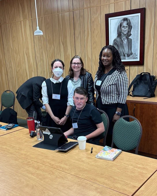 [Image: At the tenBroek Disability Law Symposium, Sam Crane, Ayesha Elaine Lewis, and Britt Belwine stand by fellow presenter Otto Lana who is seated at a conference table with his AAC device in front of him.]