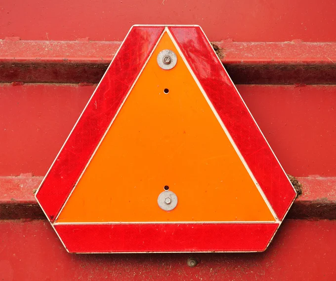 An orange triangular caution sign with a red reflective border is mounted on a textured red surface, secured with two round fasteners.