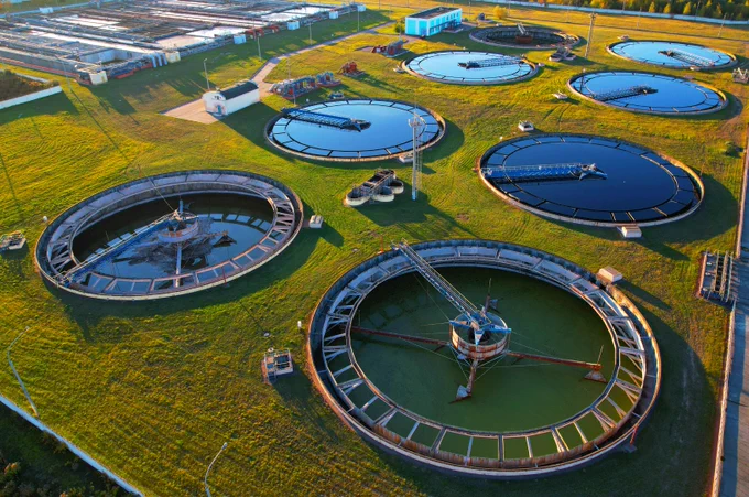 Aerial view of a wastewater treatment plant with multiple circular sedimentation tanks on green grass under sunlight.