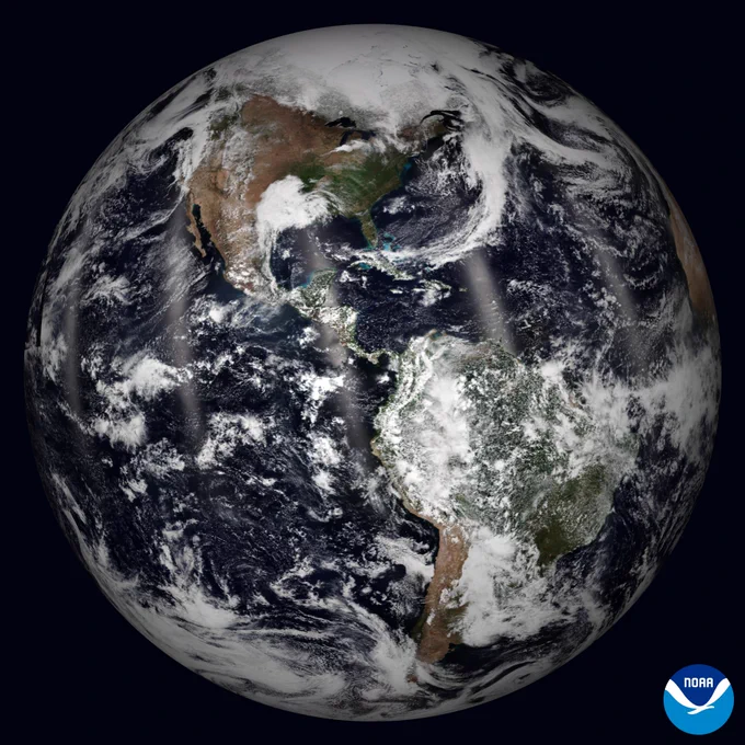 Global satellite view of Earth from April 21, 2026, centered on North and South America, with swirling white clouds against a dark background. A NOAA logo can be seen in the bottom right corner.