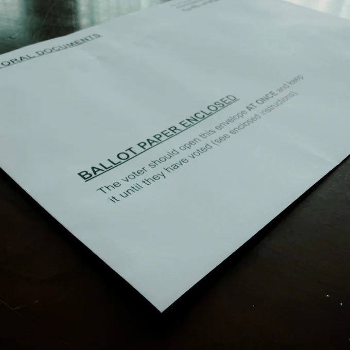White envelope labeled “Ballot Paper Enclosed” on a wooden table, with smaller printed instructions advising the voter to open it only when ready to vote.