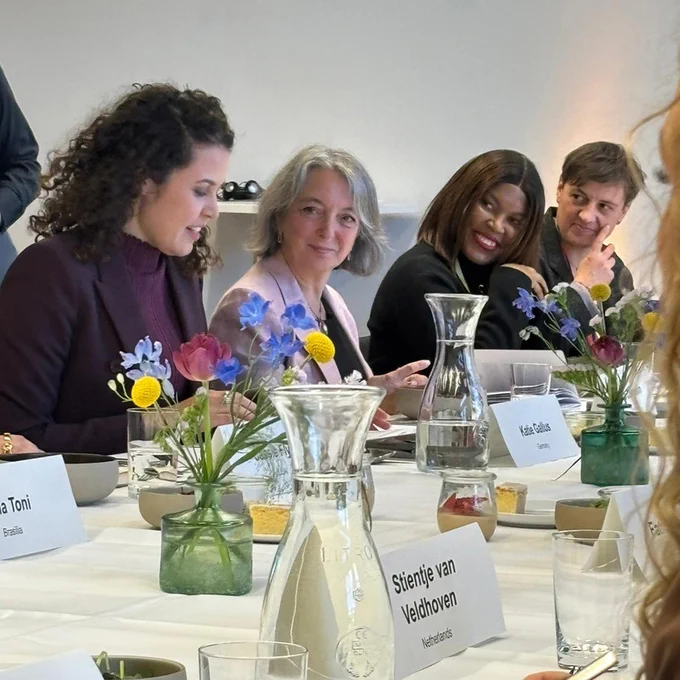 Minister Dabrusin seated alongside women representatives at a long meeting table, with small jars of colorful flowers, water carafes, notebooks, and name cards.