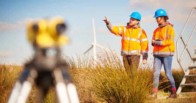 Two individuals in orange safety vests and blue helmets stand in a grassy field with wind turbines in the background. One person is pointing, and a tripod with equipment is visible in the foreground.
