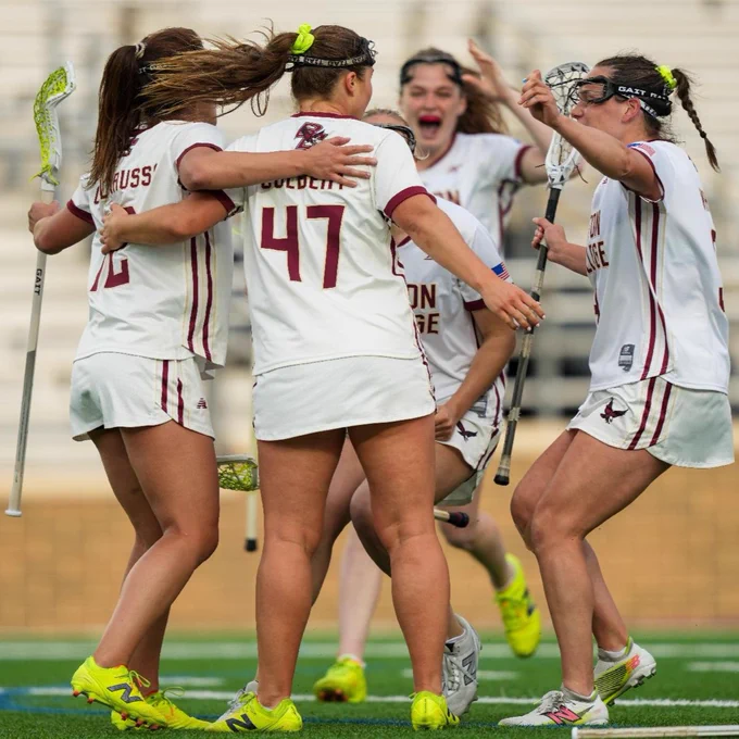 A group of Boston College lacrosse players in white and maroon uniforms celebrate on the field with lacrosse sticks, forming a huddle.