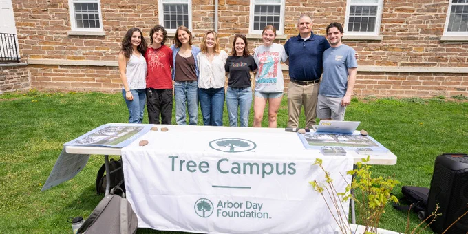 A group of people stands outside behind a table with a "Tree Campus" banner, representing the Arbor Day Foundation. They are in front of a brick building. The table displays some materials and a laptop.