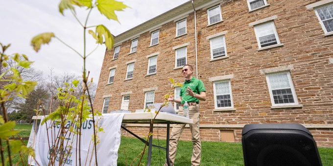 A person stands at a table with promotional materials in front of a large brick building. A white banner on the table reads "The Campus We All Call Home" and "Bucknell University." A speaker is placed on the grass nearby.