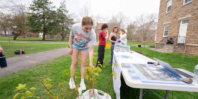 A person wearing a T-shirt bends to examine a plant placed on the ground. Nearby, a table is set up with informational materials on a college campus, and several other people are engaged in activities around it. The setting is outdoors with grass, trees, and a brick building visible in the background.