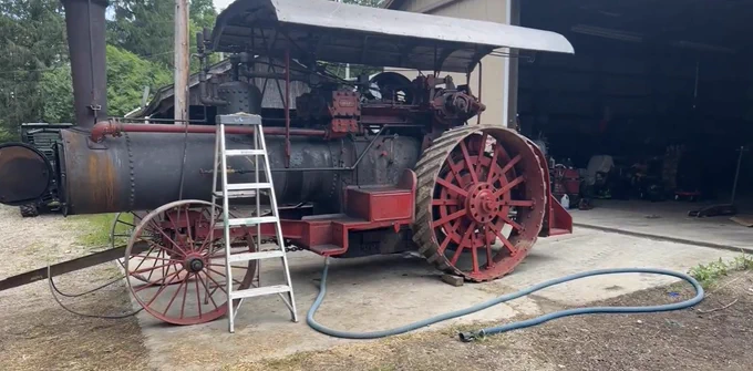 A large, vintage steam engine tractor is parked outside a garage. The tractor is weathered with a dark body and large red spoked wheels. A metal ladder leans against it, and a hose is coiled on the ground nearby. The garage is partially open, and trees are visible in the background.