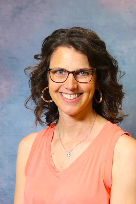 Woman with curly dark hair wearing a sleeveless coral top and hoop earrings against a soft blue-gray backdrop.