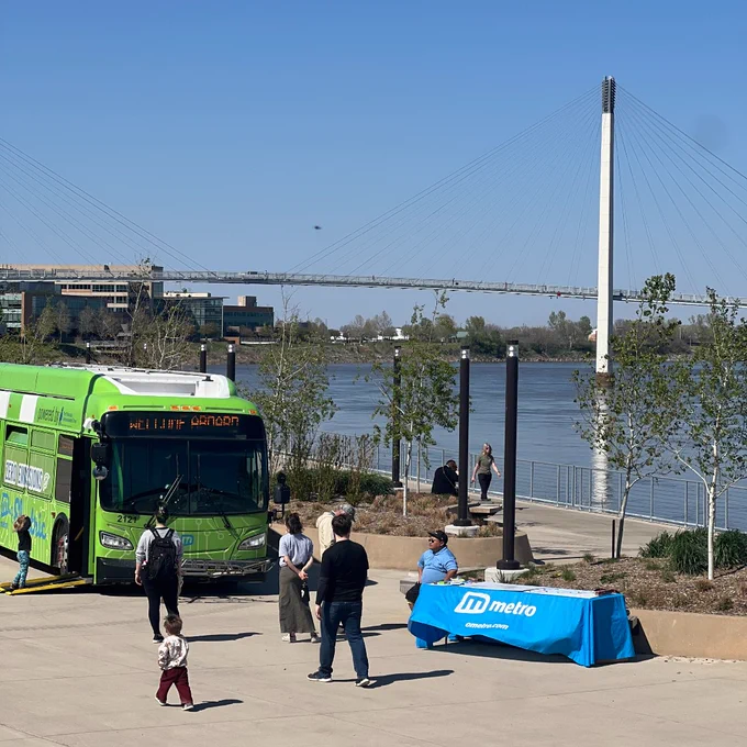 One of Metro Transit's bright green electric buses parked on the Lewis & Clark landing. There is an informational table with a bright blue table cloth that says "Metro" next to the bus. The Bob Kerrey pedestrian bridge is in the background.