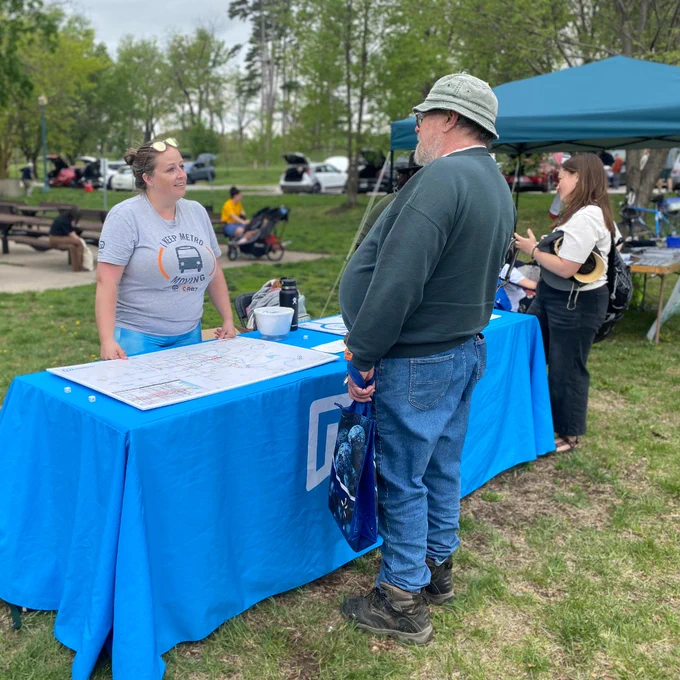 Man in a hat and green sweater talks to a woman at an outdoor booth covered with a blue tablecloth during a park event.