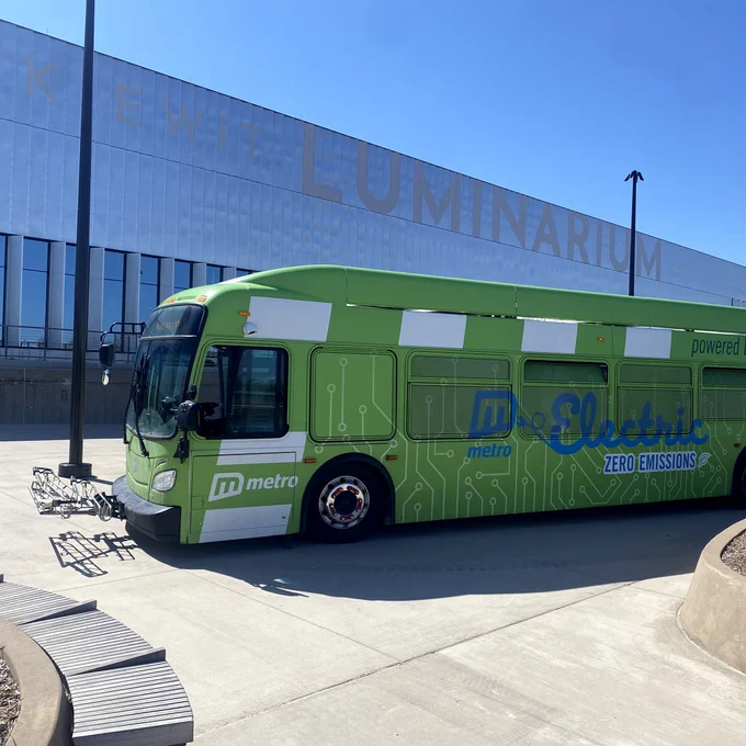 Green electric metro bus parked outside a modern building with 'LUMINARIUM' signage on a clear sunny day. The side of the bus says "Electric, zero emissions"