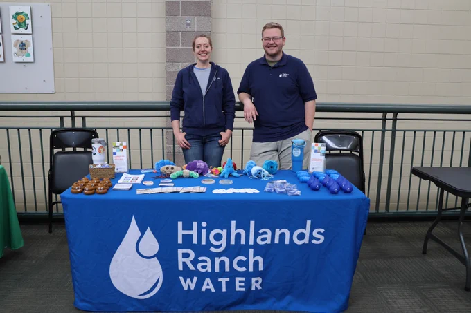 Woman and man standing behind Highlands Ranch Water's blue event booth, smiling, posing for a photo in a school hallway.
