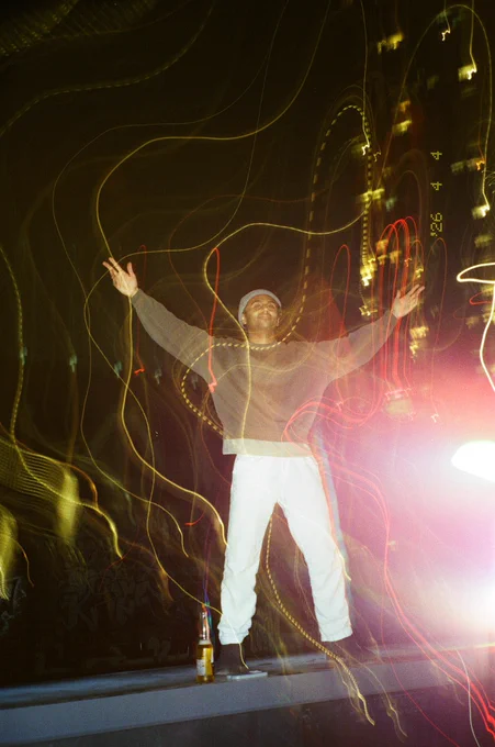 man standing on the edge of a rooftop, arm outstretched above him with heavy light leaks and light trails shot on film.