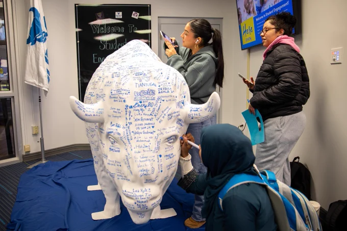 Three U B students adding their signatures to a large buffalo statue in the Student Engagement office.