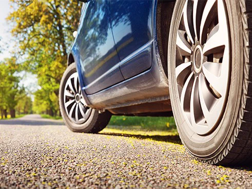 A blue car parked on a scenic, sunlit road lined with trees. The focus is on the car’s wheels and the texture of the asphalt.