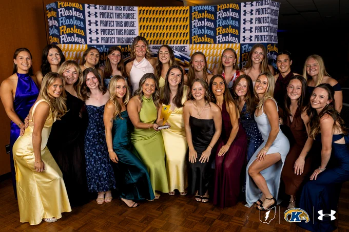 A group of twenty individuals in formal attire poses for a photo at an event. They are standing on a wooden floor, and the backdrop displays the words "Protect This House" repeatedly, along with logos for Under Armour and Kent State University. One person holds a trophy at the center.