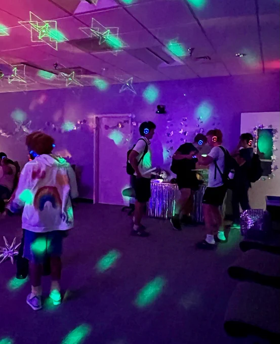 A group of people are enjoying a silent disco party in a dimly lit room. They are wearing headphones with glowing LED lights in blue, green, and red. The room is decorated with stars on the ceiling and walls. The atmosphere is lively and colorful.