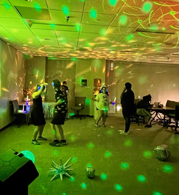 A group of people dances in a dimly lit room with colorful disco lights. The atmosphere is lively, with reflections on the walls. Some individuals are seated at a table on the side.