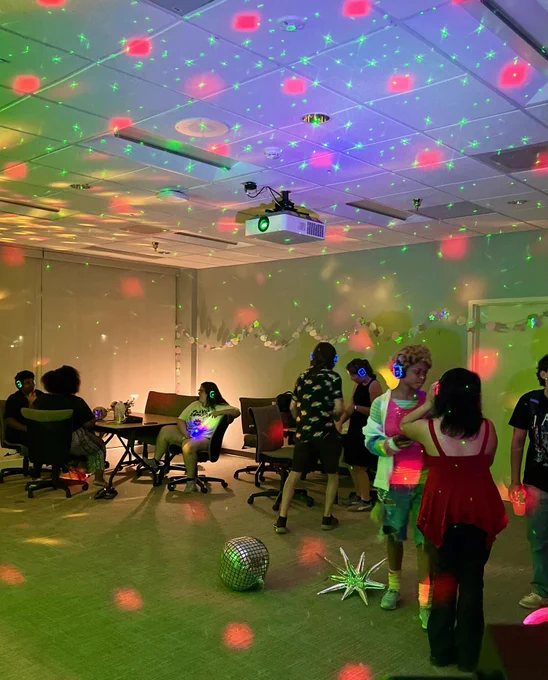 A group of people are enjoying a silent disco party in a dimly lit room. They are wearing headphones with glowing LED lights in blue, green, and red. The room is decorated with stars on the ceiling and walls. The atmosphere is lively and colorful.