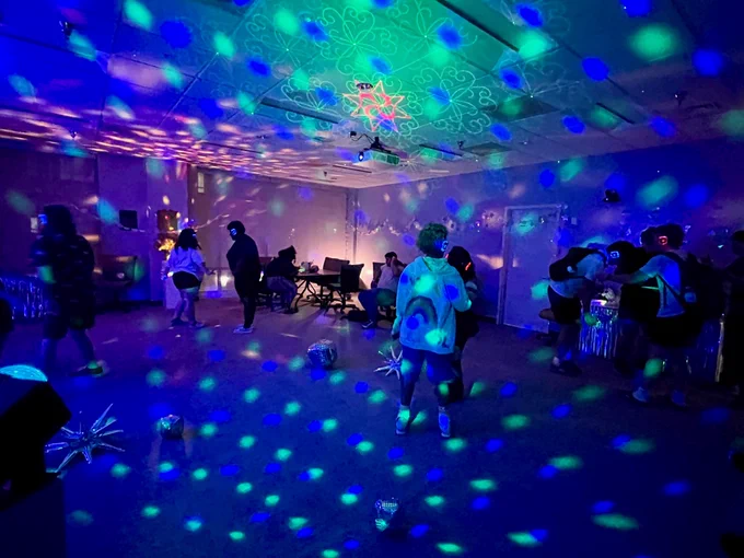 A dimly lit room features colorful, patterned lights projected on the walls and ceiling. Several people are dispersed throughout the space, some near tables and chairs in the background. The room has a festive and relaxed atmosphere.