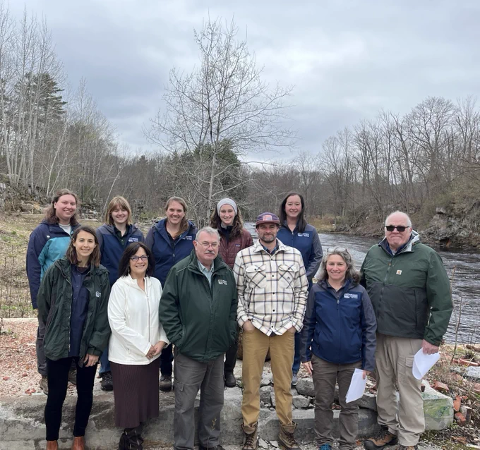 Group photo of eleven people standing on a concrete slab beside a river.