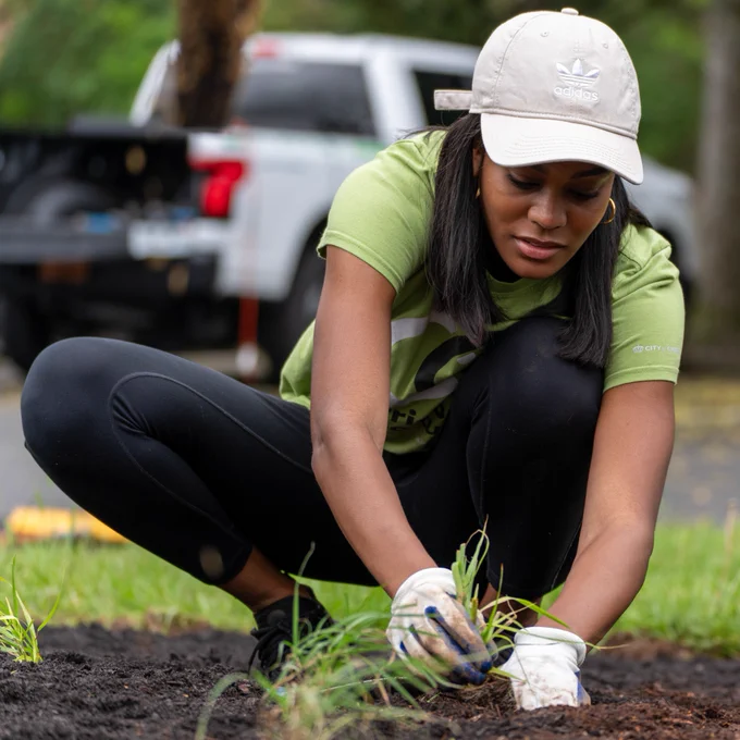 A person wearing an Adidas cap and a green t-shirt is crouching and gardening. A white truck is seen parked in the background.