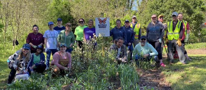 A group of people are smiling and posing outdoors in a garden area. They are standing around a sign that reads "Butterfly Highway, North Carolina Wildlife Federation." Some are wearing caps and holding gardening tools. A dog is sitting in the front. The background is filled with trees and plants.