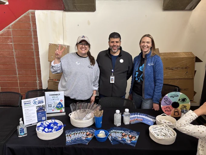 A group of three people stand behind a table displaying various informational materials and promotional items related to Charlotte Water. The table includes pamphlets, buttons, pens, and a small spinning wheel.