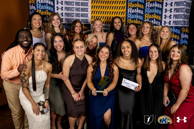 A group of people smiling and posing together in formal attire, with a backdrop featuring phrases like "Protect This House" and logos for Under Armour and Kent State University. One person holds a rectangular award plaque.