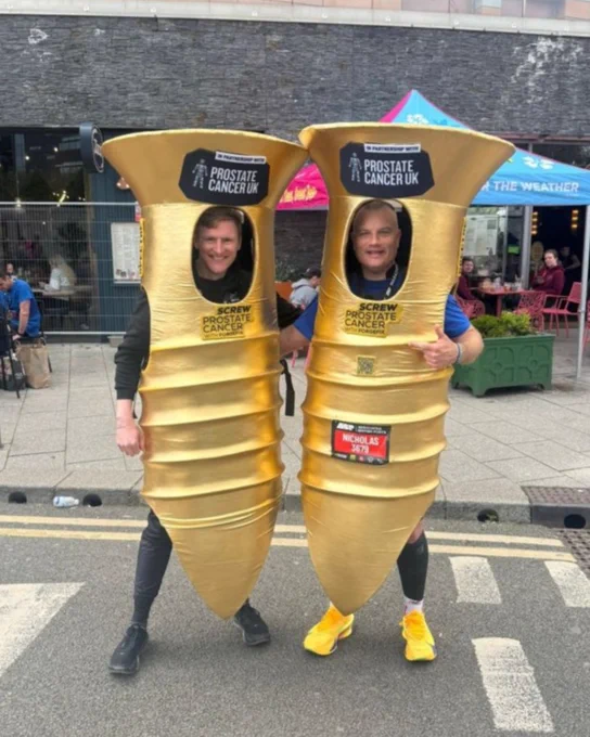 Two people are posing outdoors, dressed in large gold screw costumes with Prostate Cancer UK logos.